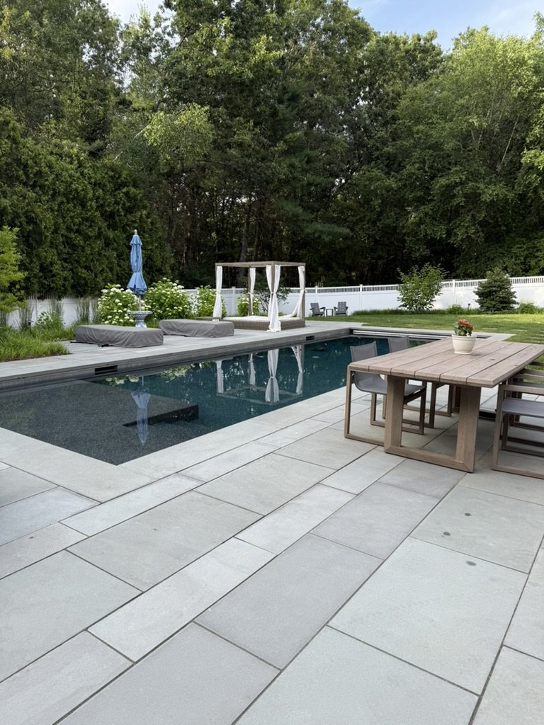 A pool with a stone patio, a wooden dining table beside it and a canopy bed at the far end, trees behind.